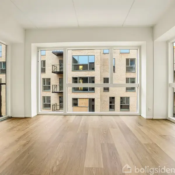 Et lyst, tomt rum med store vinduer på tre sider, der afslører udsigten til moderne lejlighedsbygninger udenfor. Gulvet er af lyst træ, der giver en varm tone. Ingen tekst.