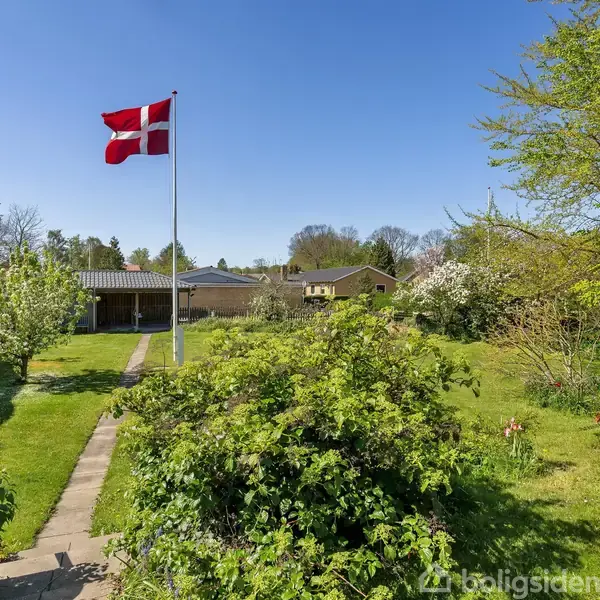Dansk flag vejrer fra en flagstang i en velholdt have. Omkranset af grønne buske og træer, med en flisebelagt sti gennem græsplænen. Klar blå himmel i baggrunden.