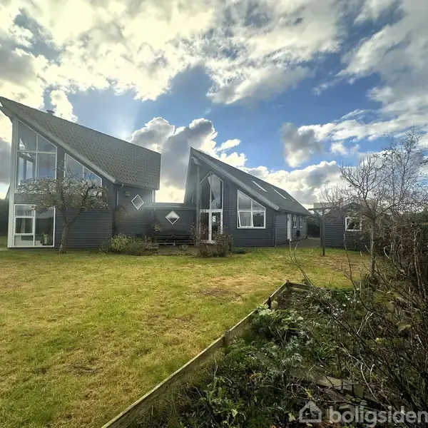 Moderne hus med skrå tage står fredeligt i en grøn have omgivet af buske og træer under en lettere skyet himmel.