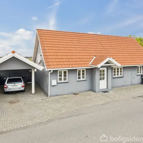 Et gråmalet hus med rødt tag står ved en vej. En sølvfarvet bil er parkeret i en carport til venstre. Baggrunden viser åbne marker.