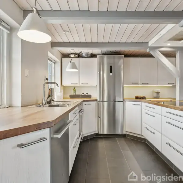 Et moderne køkken har hvide skabe, en rustfri stål-køleskab og en opvaskemaskine. En lyserød orkidé står på vindueskarmen. Rummet er lyst med store vinduer og en stilren indretning.