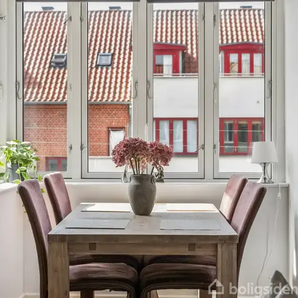 Et spisebord med fire stole står ved et stort vindue. Bordet har en vase med blomster. Udenfor ses bygninger med røde tage. Der er også en lampe og en plante.