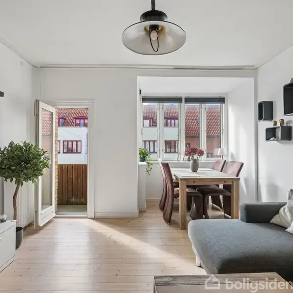 En stue med et sofaarrangement til højre, et spisebord ved vinduet og en åben altandør til venstre. Trægulve og vægmonterede hylder skaber en moderne atmosfære.