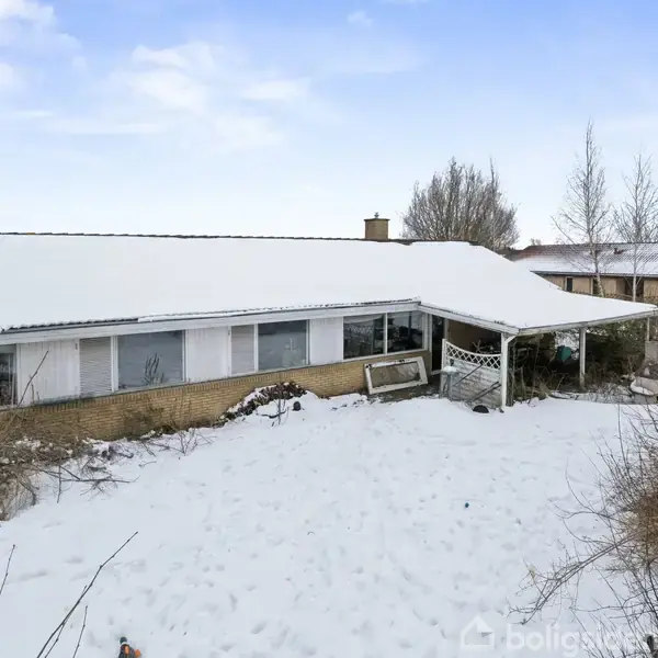Et fladt hus med sne på taget står i en snedækket have. Store vinduer vender mod kameraet, omgivet af vildtvoksende buske og træer.