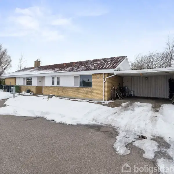 En lav, gulstenshus står stille i et snedækket område med en tilhørende carport. En parkeret bil ses til venstre på asfaltvejen, omgivet af træer.