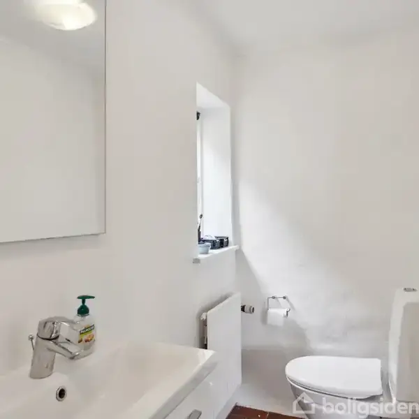 Spejl og vask står i et lyst badeværelse med en hvid væg. Håndsæbe ved vasken, toilet ved siden af, og lille vindue over en radiator. Ingen tekst står i billedet.