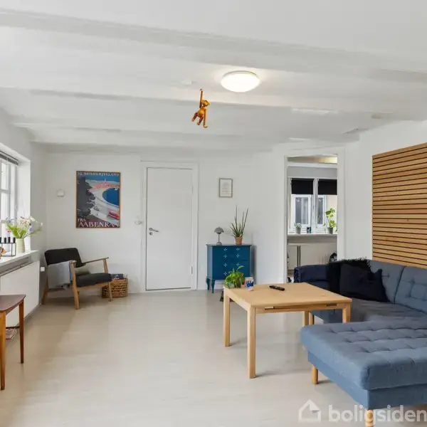 Stue med blå hjørnesofa ved sofabord, dekoreret med planter og kunst på væggen. Fladskærms-TV på venstre side. Vinduer giver naturligt lys. Lyst og moderne interiør.