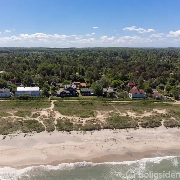 Huse langs en kyst med sandstrand og stigende bølger. Husene er omgivet af frodige grønne skove under en blå himmel med spredte skyer.