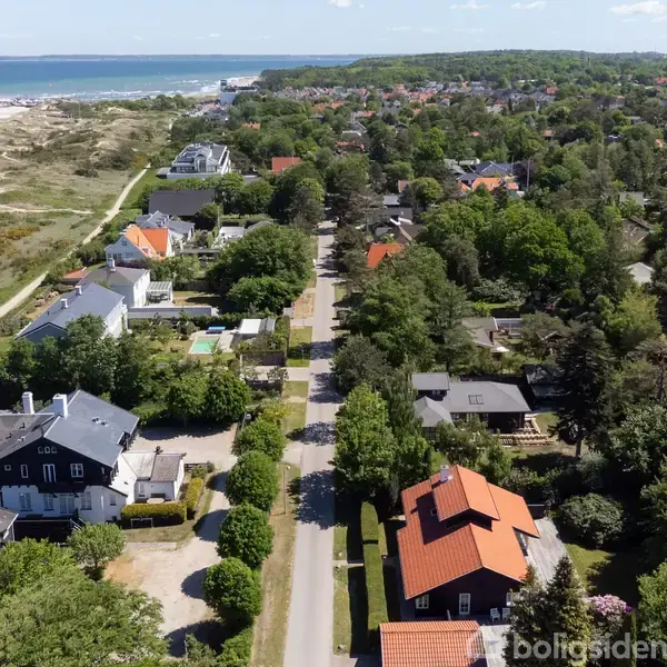 En kystnær by omgivet af grønne træer med huse langs en vej. Havet ses til venstre, mens huse med røde tage ligger blandt træerne på begge sider af vejen.