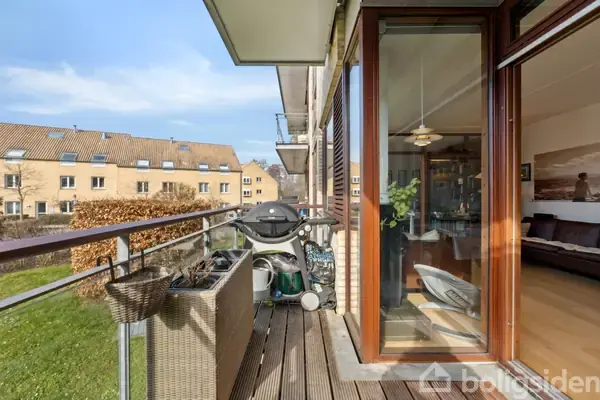 En moderne altan med brune træplanker har en grill og en flettet plantekasse. Indenfor ses en stue med trægulv, sofabord og et stort vægmaleri. Udsigt til rækkehuse.