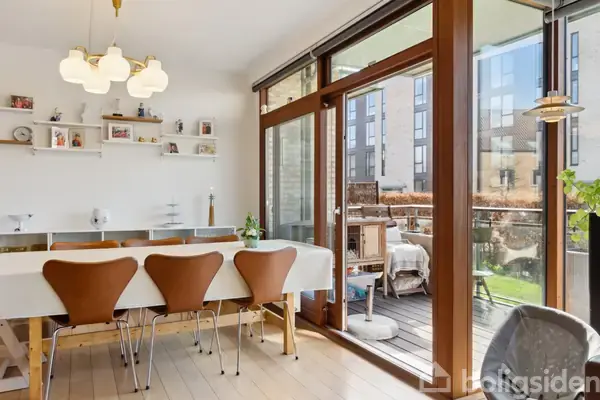 Et spisebord dækket med en hvid dug står foran tre brune stole, placeret ved siden af en glasdør, der leder ud til en balkon i bymiljø med lejlighedsbygninger.