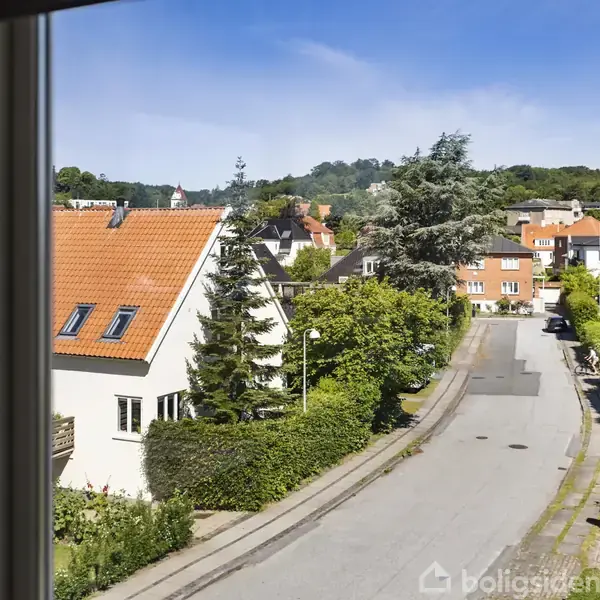 Udsigt fra et vindue viser en stille villavej med huse og haver. Det er en solrig dag, omgivelserne er frodige, og der er grønne træer i baggrunden.