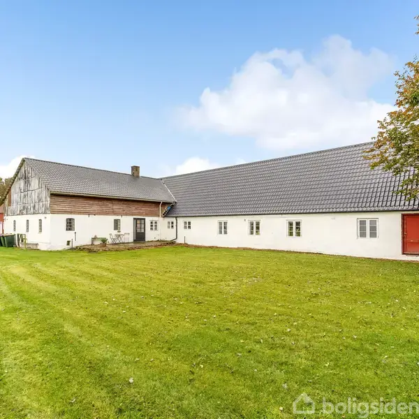 En hvid gårdbygning med rødt tag står stille i en grøn have, omgivet af træer under en blå himmel med hvide skyer.