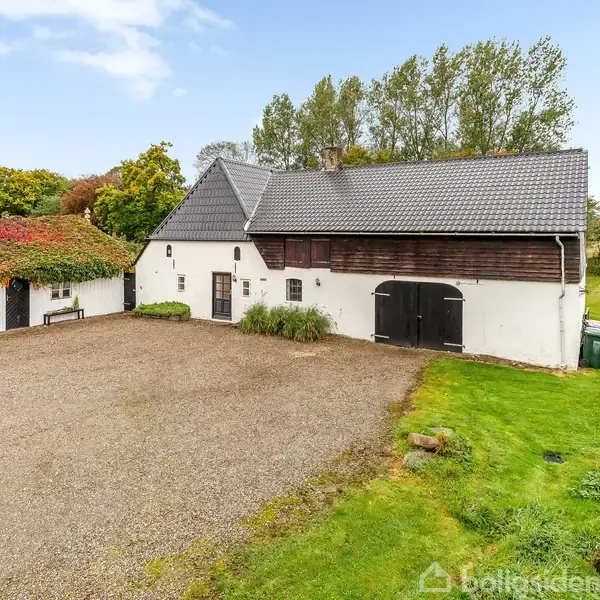 Hvidt hus med sort tag og garagedøre står på en grusbelagt plads. Der er grønne plæner og træer i baggrunden, mens en del af taget er dækket af rødt løv.