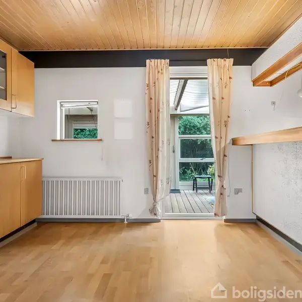 Trægulv og træloft pryder et rum med lyse træskabe. Et vindue med gardiner åbner ud til en terrasse med udsigt til grønne omgivelser. radiator monteret på væggen.