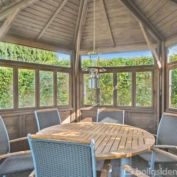 Rundt bord omgivet af seks stole står inde i en trækonstruktion med mange vinduer. Lysekrone hænger over bordet. Omgivet af grønne buske og træer.