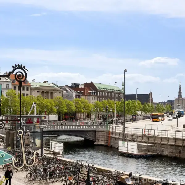 Et bybillede viser en bro over en kanal med cykler parkeret forrest. Folk går langs gaden, hvor træer står ved siden af bygninger. Skilt: "GS RESTAURANT".