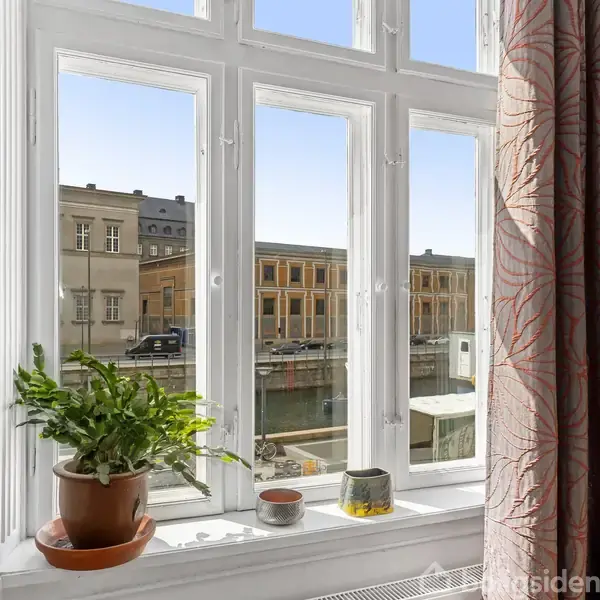 Et vindue viser en udsigt til en kanal omkranset af bygninger. En plante og to lysestager står på vindueskarmen. En lilla mønstret gardin hænger til højre.