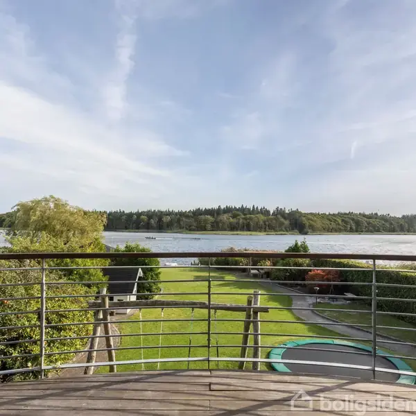 En altan med metalgelænder præsenterer udsigt over en velplejet have, der gradvist skråner ned mod en sø med skovklædte bredder i baggrunden. Trampolin i haven.