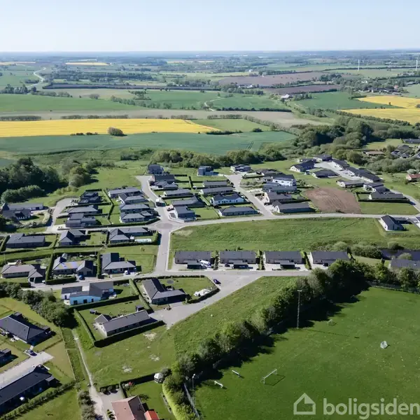 Boligområde set fra luften viser moderne huse omgivet af grønne marker og træer. Veje snor sig gennem bebyggelsen, mens åbne landbrugsområder og skove strækker sig i horisonten.