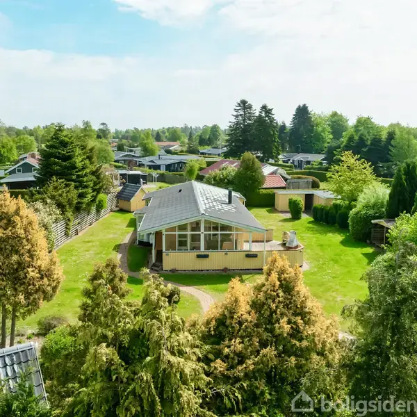 Et hus ligger centralt omgivet af en grøn have med træer og buske. En række af lignende huse strækker sig i baggrunden under en blå himmel med få skyer.