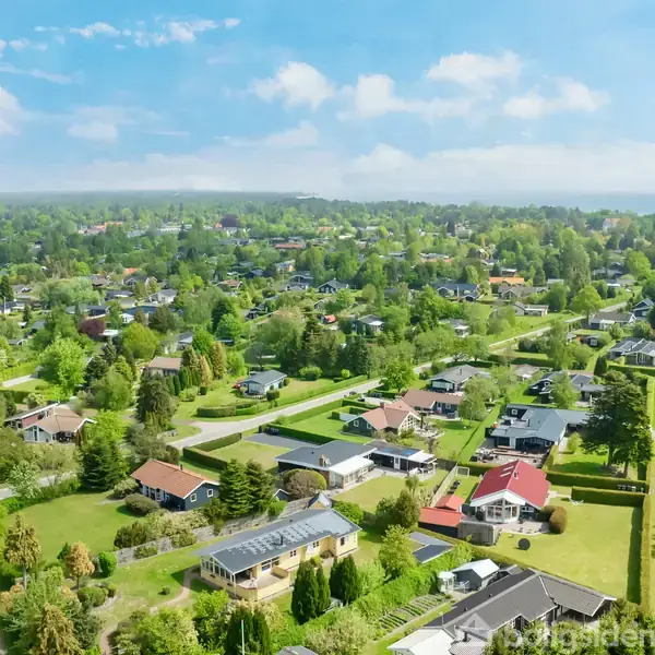 En beboelsesbygning set ovenfra, omgivet af grønne haver og veje. Bygningerne ligger i et frodigt, skovrigt område under en klar blå himmel med spredte skyer.