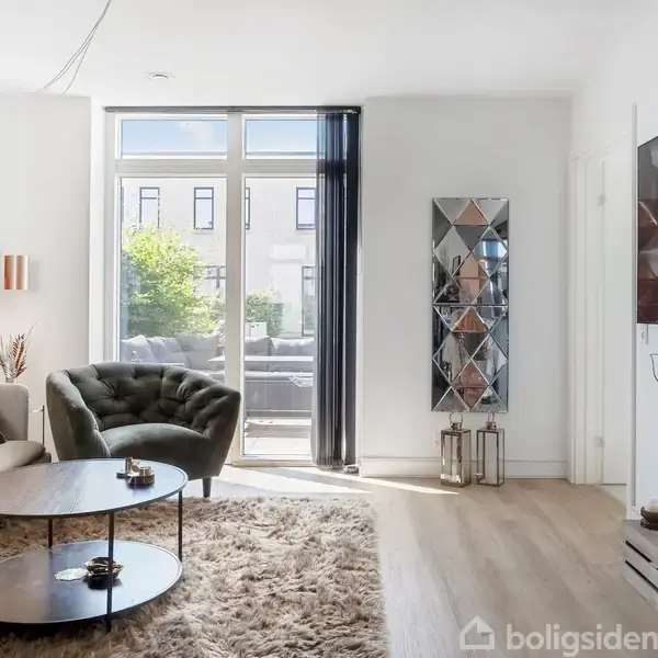 Stue med sofa og lænestol ved siden af to runde borde. Et stort vindue med udsigt til en terrasse. Et TV hænger på væggen over et arrangement med dekorationer.