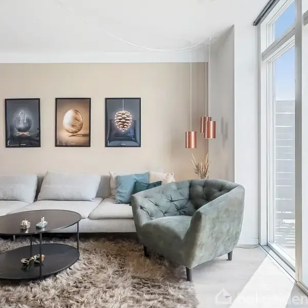 Sofa med farverige puder står i moderne stue ved siden af et grønt, polstret lænestol. Runde, sorte borde på en fluffy tæppe; store vinduer giver lysindfald. Vægkunst hænger bagved.