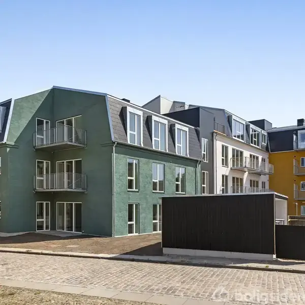 Flere fleretagers bygninger står stille langs en brostensbelagt vej. Bygningerne er malet i forskellige farver, såsom grøn og gul, og har små altaner og mange vinduer.