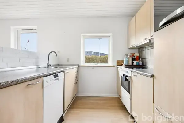 Et køkken med lyse træskabe og sort bordplade. En opvaskemaskine og ovn blandt apparaterne. Et lille vindue viser udsigt til hække og tag. Lyse trægulve.