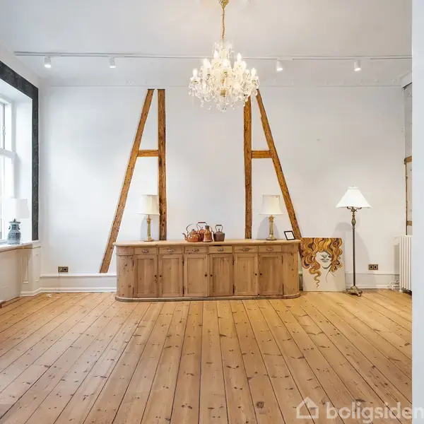 Et rum med et træskab under arkitektoniske bjælker. En lysekrone hænger fra loftet. Et vindue til venstre og en gulvlampe til højre tilføjer lys.