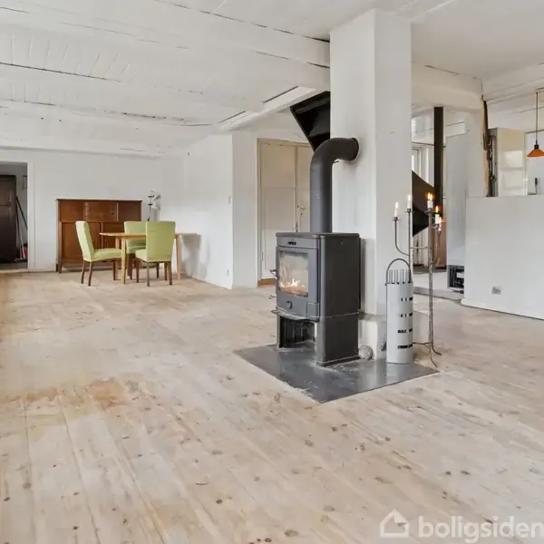 En brændeovn står midt i en rummelig stue med lyst trægulv, hvor der i baggrunden ses et spisebord med grønne stole. Der er flere vinduer, som giver rummet lys.