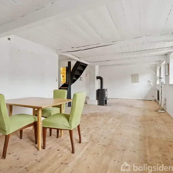 Et bord med to grønne stole står i et lyst, rummeligt rum med trægulv og hvidmalet træloft; trapper og brændeovn ses i baggrunden.