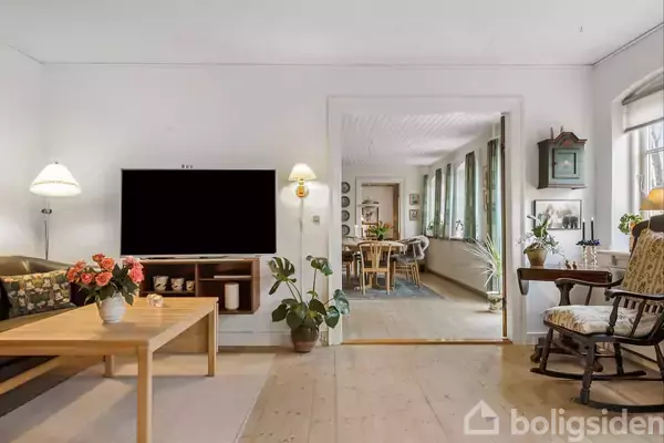 En stue med en fladskærms-tv på væggen og en træbord med blomster. Rummet er indrettet med stole og lamper, og der er udsigt til en spisestue i baggrunden.