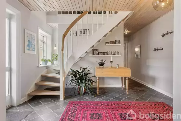 En vindeltrappe fører opad i en lys entré, hvor en grøn plante står på trægulvet. En trækommode og hylde med dekorationer i baggrunden. En rød orientalsk tæppe ligger på flisegulvet.