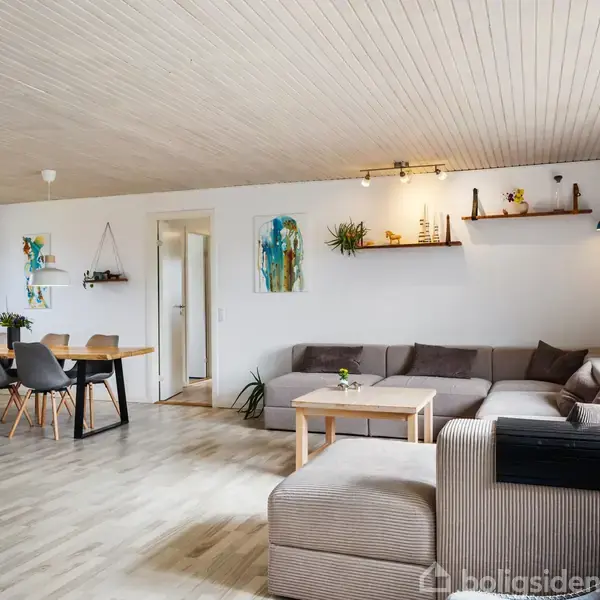 Stue med en grå hjørnesofa mod væggen, bordet står foran. Til venstre er et spisebord med grå stole. Billeder og planter pynter væggene. Store vinduer til højre.