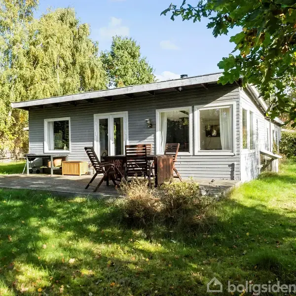 Et gråt træhus står stille i en solrig have. Foran huset er et terrassedæk med bord og stole. Baggrunden domineres af frodig vegetation og træer.