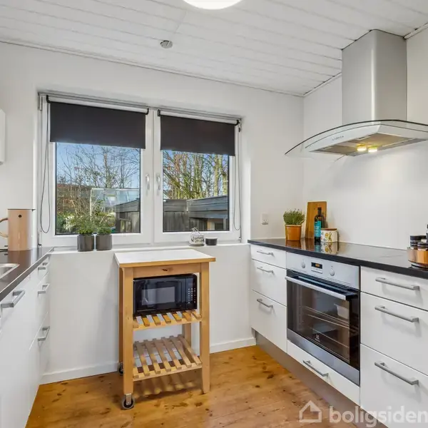 Et lyst, hvidt køkken med trælaminatgulv indeholder en ovn, emhætte, vask og en rullebord med mikrobølgeovn. To vinduer med sorte rullegardiner giver udsigt til træer udenfor.