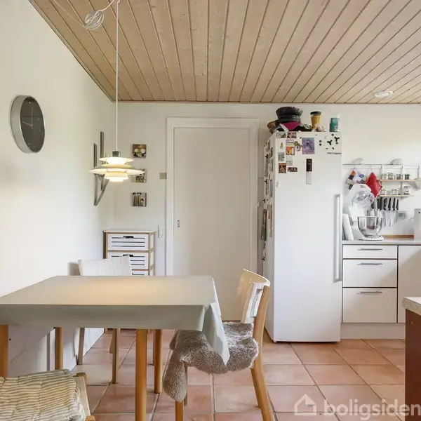 Et køkken med spisebord og fire stole, hvoraf én har et pelsklæde. Et køleskab står ved siden af skabe og hylder med køkkenartikler. Lyse vægge og trægulv.