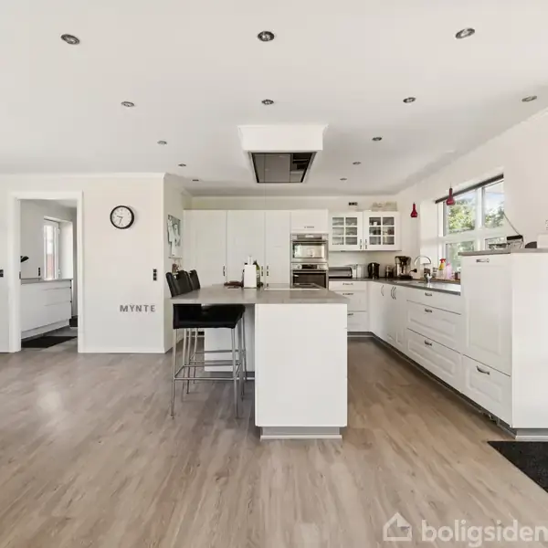 Køkken i moderne design med hvidt møblement og køkkenø. Spisestole står ved øen. Rummelige, lyse vinduer giver naturligt lys. Væggen har tekst: "MYNTE". Vægur hænger ved indgang.