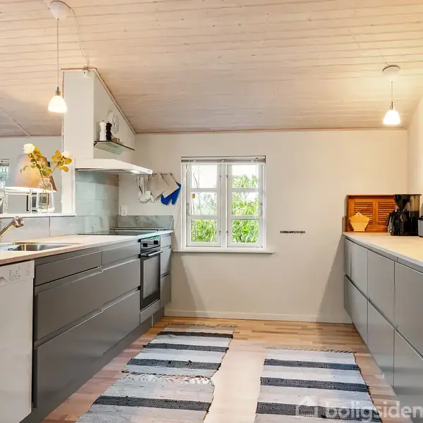 Et moderne køkken med grå skabe har en opvaskemaskine og indbygget ovn. Der hænger lamper over en lys træbordplade. Et lille vindue lader naturligt lys komme ind.
