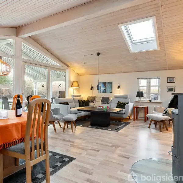 Et lyst, rummeligt stueområde med spisebord dækket af orange dug, omgivet af træstole. I baggrunden er der en sofagruppe med moderne lamper. Store vinduer og et skråt loft med ovenlysvindue tillader naturligt lys at strømme ind. En brændeovn til højre understreger hyggen.