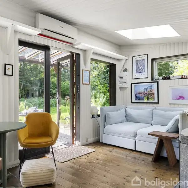 En stue med en lyseblå sofa ved siden af et vindue. En gul stol står foran en åben glasskydedør mod en grøn have. Væggene har flere kunstværker.