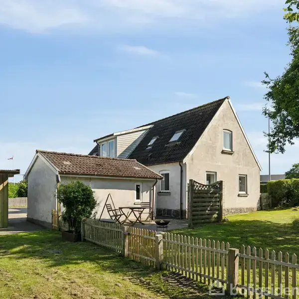 Et hvidt hus med skråt tag står stille i en solrig have med en lille terrasse foran. Træhegn omgiver græsplænen, hvor træer giver skygge.