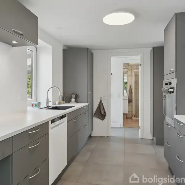 Et moderne køkken med grå skabe og hvide bordplader. En indbygget ovn og komfur findes til højre, mens en vask og opvaskemaskine er på venstre side. Axelyst overfyldes rummet.