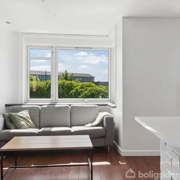 En grå sofa står mod en hvid væg ved et stort vindue med udsigt til træer og bygninger. Foran sofaen er et rektangulært sofabord. Det er solskin udenfor.