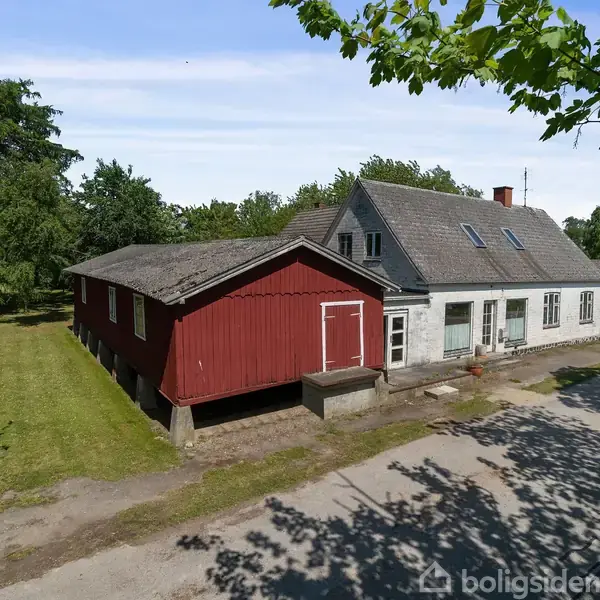 En bondegård med rødt træskur og gråt stuehus står ved en grusvej. Græsplæner med store træer omgiver bygningerne, skaber en landlig atmosfære.