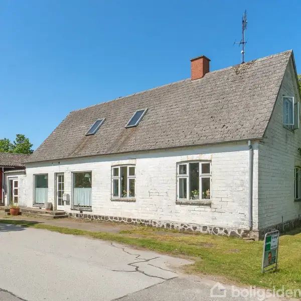 Hus med hvid facade står stille ved landevej, omgivet af grønne træer. Til venstre en mindre rød bygning. Klar blå himmel i baggrunden.