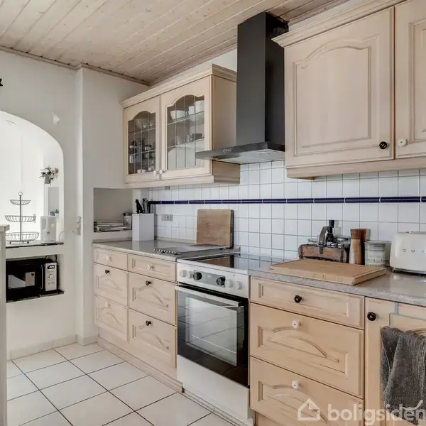 Køkken med lyse skabe og en indbygget ovn. Køkkengrej og apparater såsom en brødrister og kaffemaskine på bordet. Hvid kakler på væggen og åbning til spisestue i baggrunden.