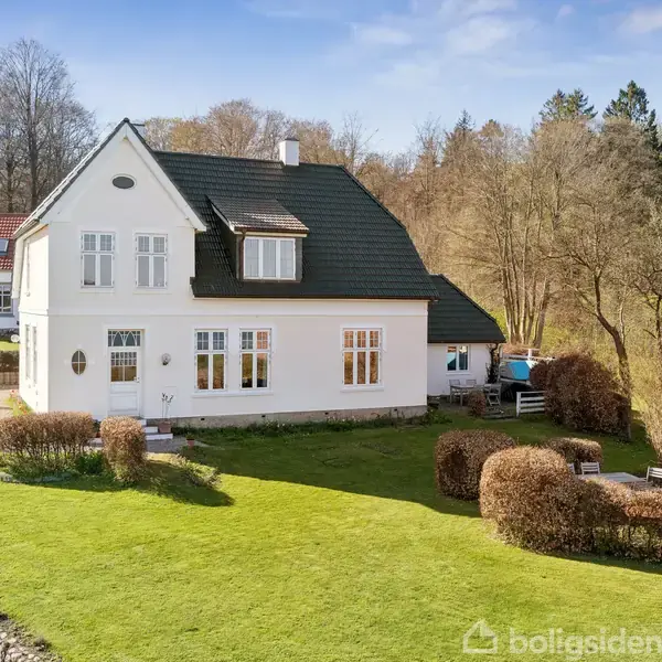 Hvidt hus med sort tag står på en velplejet græsplæne med buske. Baggrund har skov og en vej. Sollys skinner på huset, hvilket giver en lys atmosfære.